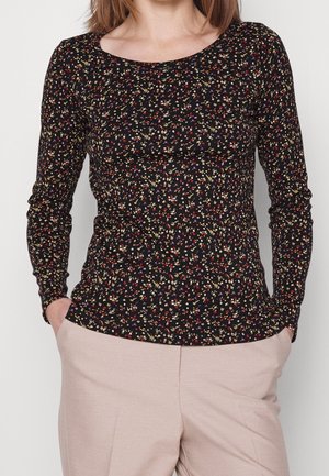Femme portant un haut noir ajusté à manches longues avec un petit imprimé floral multicolore et un pantalon beige, les mains dans les poches.
