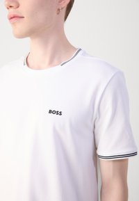 Baltas marškinėliai su juodu "BOSS" logotipu ant krūtinės. Kaklo linija ir rankovių kraštai turi juodai baltas juosteles. Trumpos rankovės, lygus tekstūras.