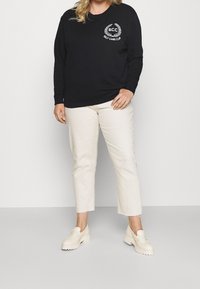 Felpa nera con logo "S.C.C" bianco e design di alloro, abbinata a pantaloni cropped crema e scarpe slip-on chunky di colore bianco sporco.