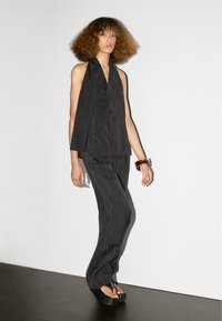 Zwart pinstripe pak met een mouwloze top met een diepe V-hals en op maat gemaakte broek. Model draagt platform sandalen en een bruine armband.