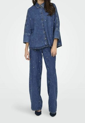 Vrouw draagt een loszittend denim shirt en bijpassende jeans met wijde pijpen, beide versierd met kleine parelachtige studs.