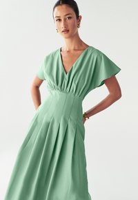 Vestido verde hecho de una tela ligera, con escote en V, mangas cortas con volantes, cintura elástica y falda plisada. Textura suave en toda la prenda.