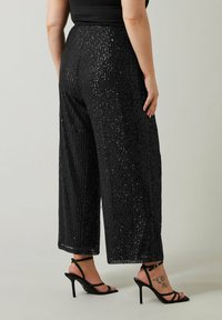 Pantalons larges noirs à sequins avec une surface texturée. Présentent une coupe ample et un ourlet fringé, associés à des sandales à talons hauts noires.