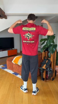 Άνδρας με κόκκινο γραφικό μπλουζάκι λυγίζοντας τα χέρια του σε σαλόνι με πορτοκαλί καναπέ, μεγάλο πράσινο φυτό, μηχάνημα κωπηλασίας και τηλεόραση.