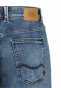 Jean en denim bleu avec coutures foncées, une poche arrière, une passant de ceinture et un patch en cuir beige portant l'inscription Camel Active sur la ceinture.