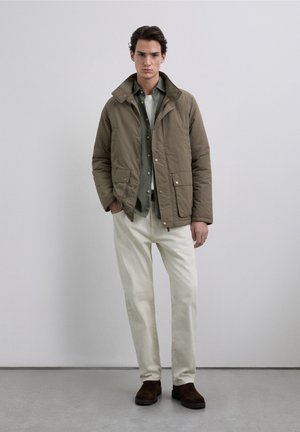 Chaqueta acolchada verde oliva con cuello alto, cierre de botones y dos bolsillos delanteros, llevada sobre una camisa clara, combinada con pantalones crema y botas marrones.