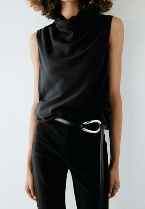 Femme portant un haut noir sans manches avec un décolleté drapé et un pantalon noir maintenu par une ceinture à boucle argentée distinctive.