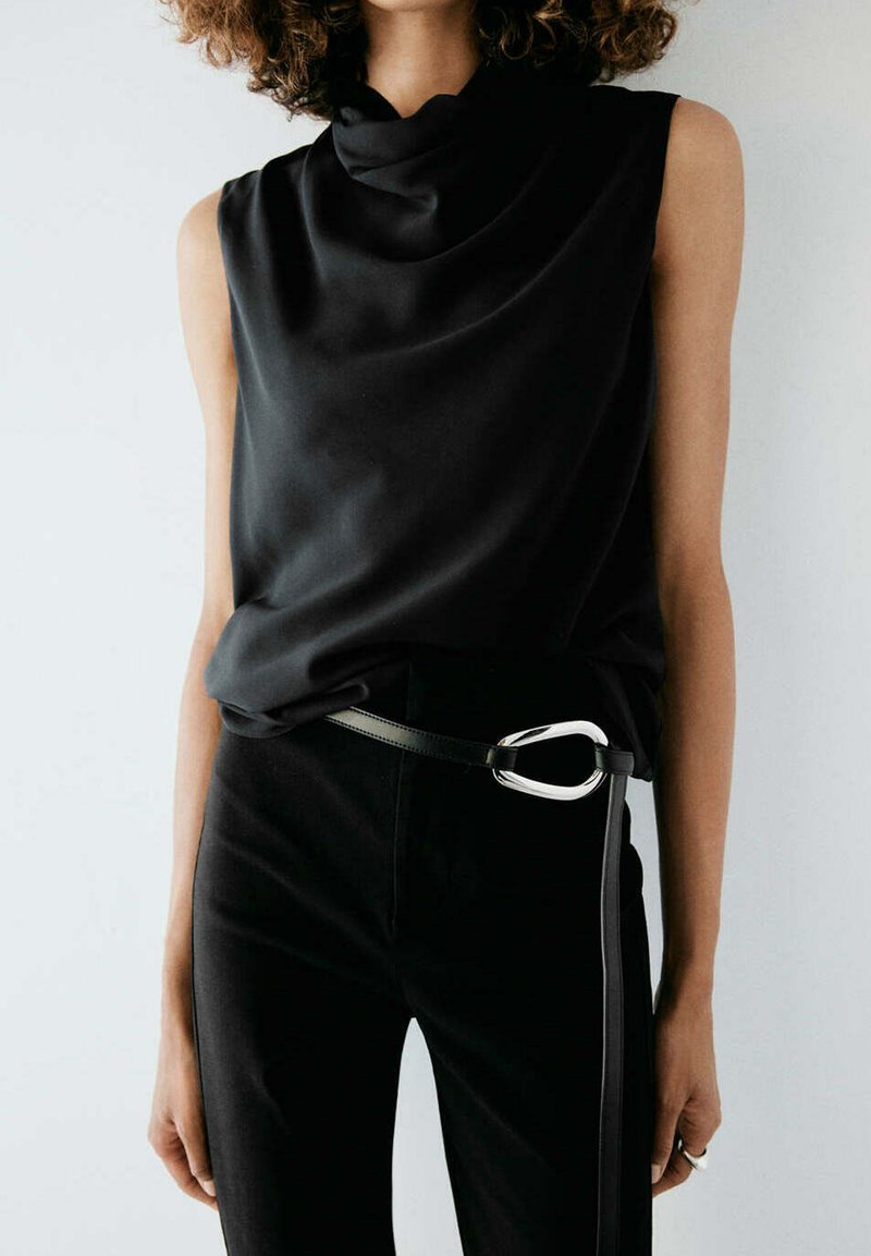 Femme portant un haut noir sans manches avec un décolleté drapé et un pantalon noir maintenu par une ceinture à boucle argentée distinctive.