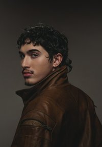 Braune Lederjacke mit hohem Kragen, Reißverschlusstasche an der Schulter, grober Textur und figurbetonter Form, getragen von einer Person in Seitenansicht.