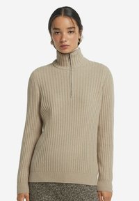 Beige ribbesweater med høj krave og lynlås foran. Lavet af blødt stof, med lange ærmer og tætsiddende design.