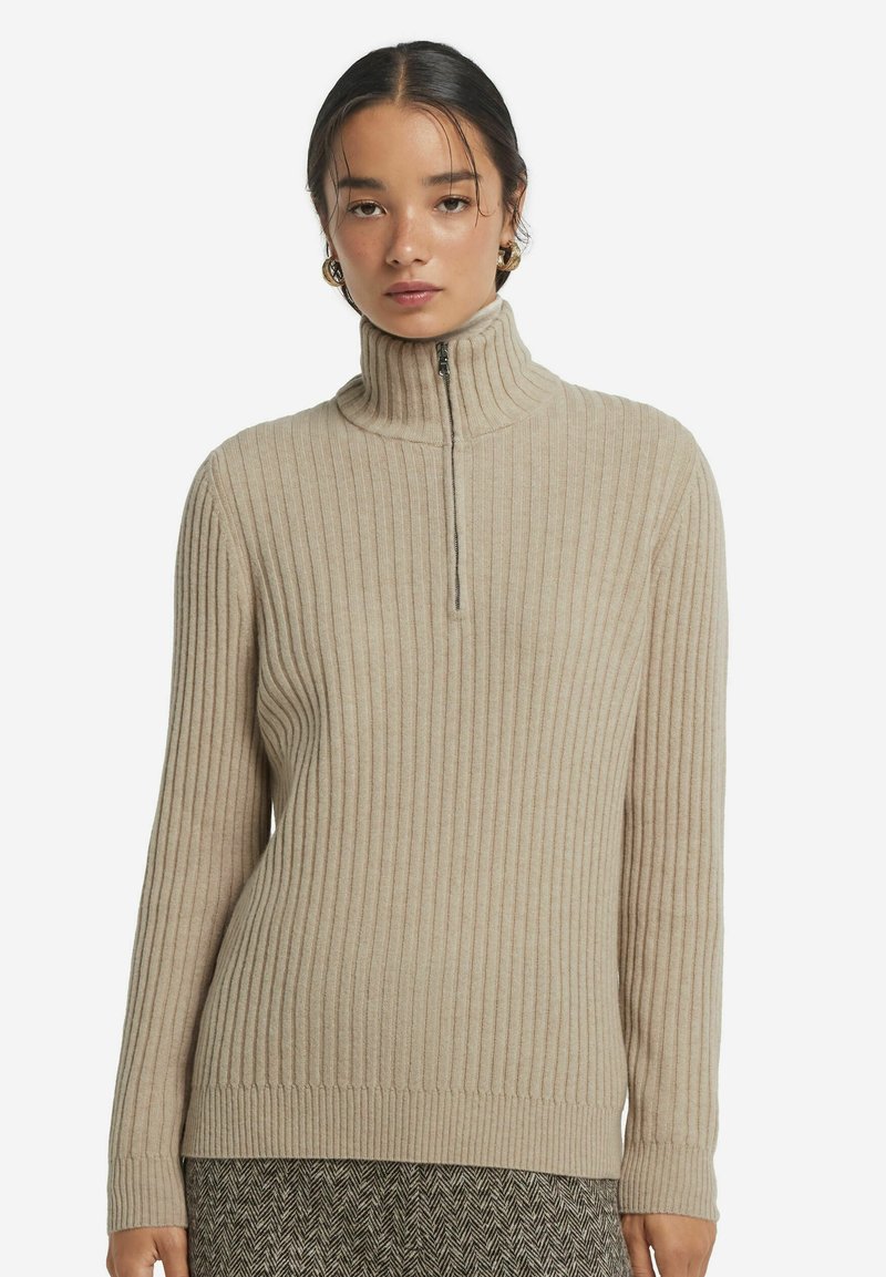 Beige ribbesweater med høj krave og lynlås foran. Lavet af blødt stof, med lange ærmer og tætsiddende design.