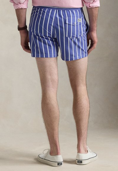 Mėlyni dryžuoti maudymosi šortai su užpakaline kišene, elastingu liemeniu ir lengva medžiaga. Dėvimi su baltais drobiniais sportbačiais.