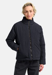 Giacca puffy di un blu navy scuro con colletto alto, chiusura con zip e dettagli di cuciture orizzontali. Presenta tasche laterali e un logo sull'orlo.