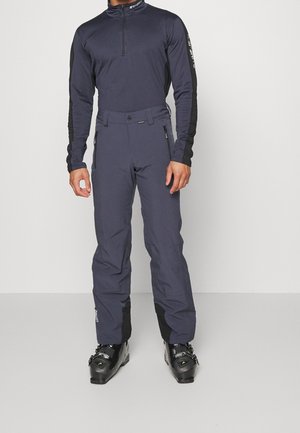 Man draagt donkergrijze skibroek en bijpassende zip-up longsleeve met zwarte skischoenen op een effen achtergrond.