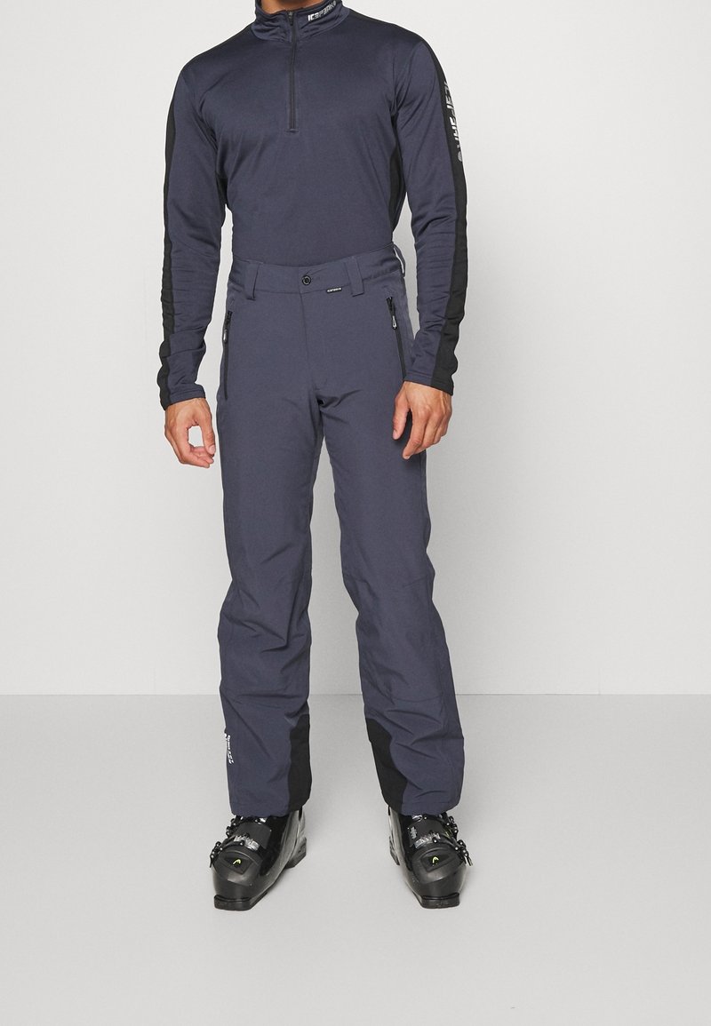 Homme portant un pantalon de ski gris foncé et un haut à manches longues zippé assorti avec des bottes de ski noires sur un fond uni.