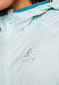 Gros plan sur une veste zippée bleu clair avec col intérieur turquoise et logo Odlo sur la poitrine, portée par une personne.