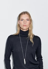 Black turtleneck sweater, soft texture, fitted design. Long silver pendant necklace, oval shape, hanging on a thin chain. Plain background.