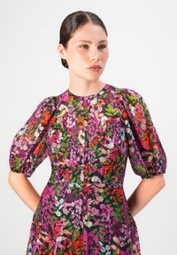 Vrouw met donker haar in een lage bun, gekleed in een kleurrijke bloemenjurk met pofmouwen, die iets opzij kijkt tegen een effen achtergrond.