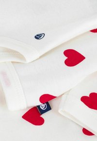 Tessuto bianco con motivi a cuori rossi, con un'etichetta logo blu visibile. Il design presenta bordi a coste e una consistenza morbida.
