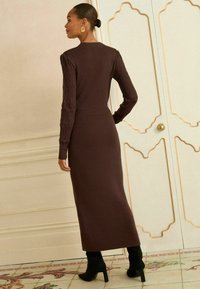 Abito a maglia marrone a maniche lunghe con maniche texturizzate, silhouette aderente e lunghezza midi. Indossato con stivaletti neri con tacco.