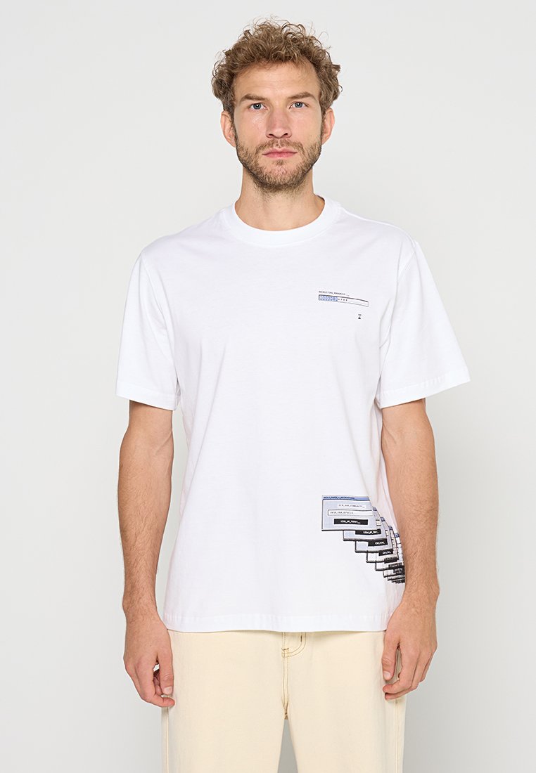 Mann trägt ein weißes T-Shirt mit kurzen Ärmeln, auf dem sich nahe der linken Brust ein kleines schwarzes und blaues grafisches Textmuster sowie grafische Tags in der Nähe der rechten Hüfte befinden.