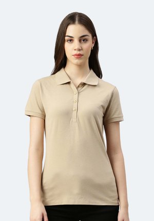 Jeune femme aux longs cheveux bruns portant un polo beige à manches courtes, debout devant un fond clair uni.