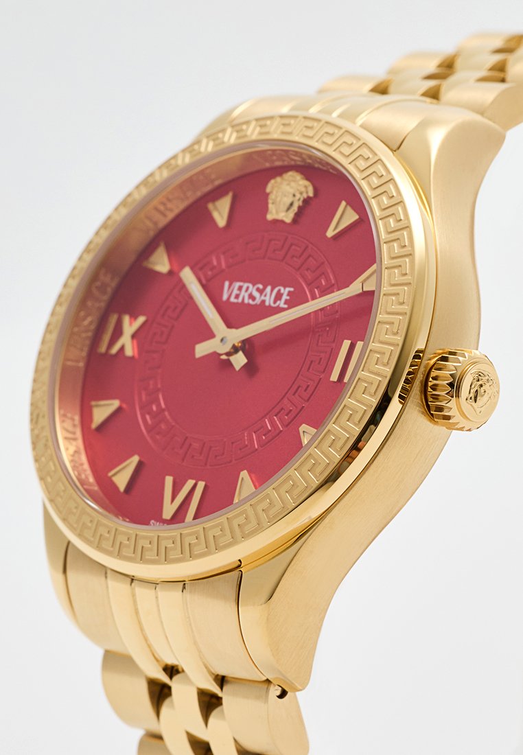 Reloj de tono dorado con una esfera roja, números romanos dorados, patrón intrincado en el bisel y un logo de león a las 12 en punto. La correa de metal añade textura.