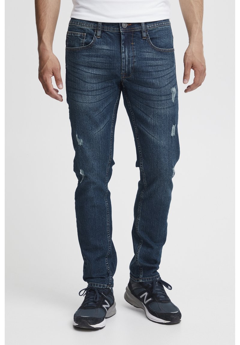 Jeans de mezclilla azul oscuro con ajuste slim, que presentan zonas desgastadas y un ligero desvanecimiento. Diseño clásico de cinco bolsillos con cremallera.