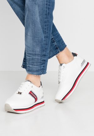 Personne portant un jean bleu et des baskets blanches avec des accents rouges, noirs et argentés, debout sur un sol lisse gris clair.