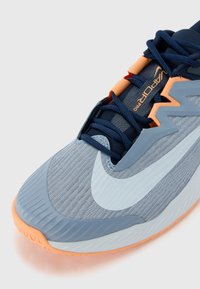 Hellblaue Sportschuhe mit einer strukturierten Mesh-Oberfläche, orangen Akzenten und einem kontrastierenden weißen Swoosh-Logo auf einer abgerundeten Sohle.