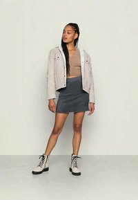 Casaco curto cinza claro, blusa castanha, saia de jeans ajustada e botins bejes com atacadores pretos e solas de borracha, de pé contra um fundo neutro.