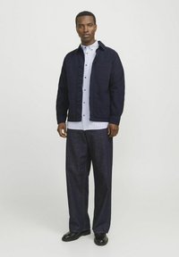 Navy blue jacket with button closure and front pockets, striped shirt beneath, paired with dark denim trousers and black shoes.