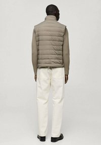 Vestă puffer bej cu un design cu matlasare orizontală, purtată peste un pulover de culoare deschisă și asortată cu blugi albi și pantofi negri.