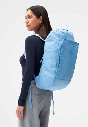 Frau mit langen dunklen Haaren, die ein marineblaues Oberteil und graue Hosen trägt, trägt einen großen hellblauen Rucksack auf dem Rücken und schaut nach links.