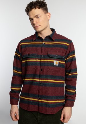 Camisa de franela a cuadros rojos con rayas amarillas y azules, de manga larga, con dos bolsillos en el pecho y cierre de botones. La tela tiene un acabado texturizado.