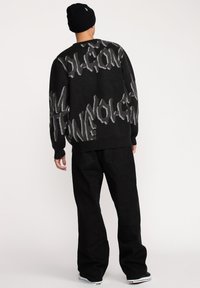 Pull noir avec motif graphique en texte gris, manches longues et col rond. Associé à un pantalon noir à jambes droites et un bonnet noir.