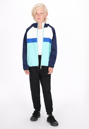 Giacca con cappuccio blu chiaro e blu navy con strisce bianche e blu; abbinata a pantaloni neri e sneakers nere. Design minimalista e vestibilità.