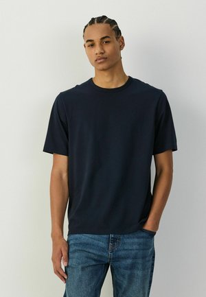 Young man with braided hair wearing a plain black t-shirt and blue jeans, standing with one hand in pocket against a light wall.