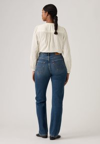 Γυναίκα με πλεκτά μαλλιά φοράει λευκό μακρυμάνικο μπλουζάκι με υφή, στερεωμένο μέσα σε ψηλόμεσα μπλε τζιν, στέκεται με την πλάτη προς τα πίσω σε ουδέτερο φόντο.