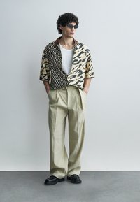 Uomo che indossa pantaloni beige larghi, scarpe nere, camicia con motivo metà a quadri e metà animalier, maglietta bianca sotto e occhiali da sole neri.