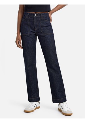 Model draagt donkerblauwe high-waist rechte jeans met opgestikte zakken en witte sneakers met zwarte strepen.