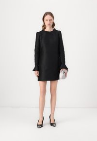 Vestito mini nero con texture, maniche lunghe e polsini svasati, abbinato a una pochette argento e tacchi neri con accenti metallici.