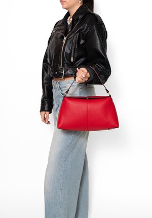 Person in black leather jacket and light blue jeans holding a red leather handbag by its short strap at their side.