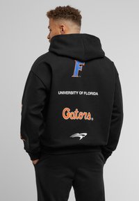 Sudadera negra con bolsillo tipo canguro. Presenta una "F" en azul, "UNIVERSIDAD DE FLORIDA" en blanco y "Gators" en naranja en la parte posterior.