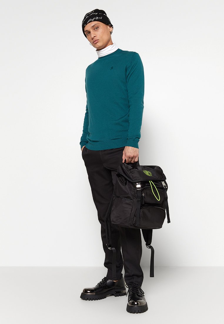 Turkusowy sweter, czarne spodnie, błyszczące czarne buty oraz czarny plecak z zielonymi akcentami. Ubranie ma okrągły dekolt i czapkę beanie.