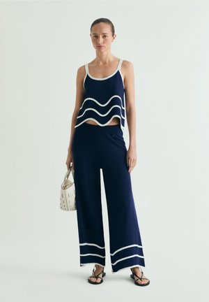 Woman standing wearing navy blue sleeveless top and wide-leg pants with white wavy trim, holding a white handbag and wearing black sandals.
