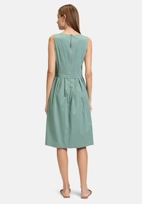 Grünes ärmelloses Kleid mit tailliertem Schnitt, ausgestelltem Rock und einem Reißverschluss auf der Rückseite. Glattes Gewebe mit einem sanften Glanz.