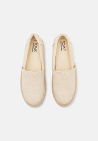 Scarpe slip-on beige con tomaia in tessuto testurizzato, scollo elasticizzato a V e suola in juta stile espadrillle di colore beige. Gli interni presentano il marchio TOMS.