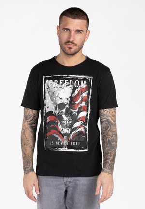 Man met kort haar en tatoeages draagt een zwart T-shirt met schedel en Amerikaanse vlag met de tekst "VRIJHEID IS NOOIT GRATIS," staand tegen een witte achtergrond.