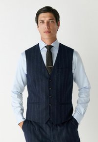 Marineblauwe pinstripe gilet van textuurstof, met vijf knopen, zijzakken, en gecombineerd met een lichtblauw overhemdd en een gestreepte stropdas.